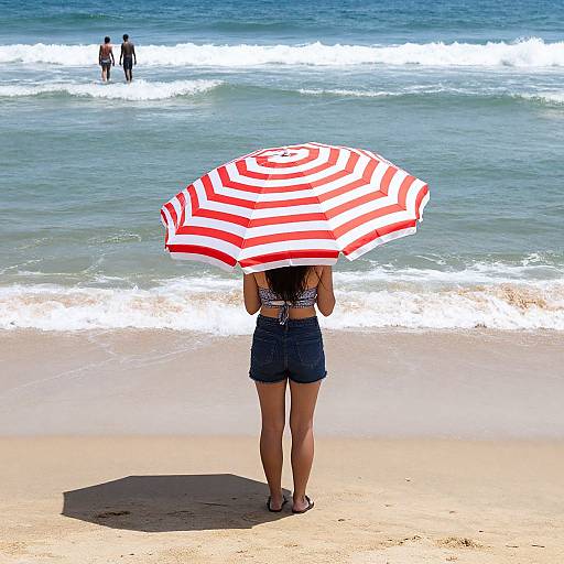 Photograph of a woman with dark hair, wearing a white crop top and denim shorts, standing on a beach under a red-and-white striped umbrella,