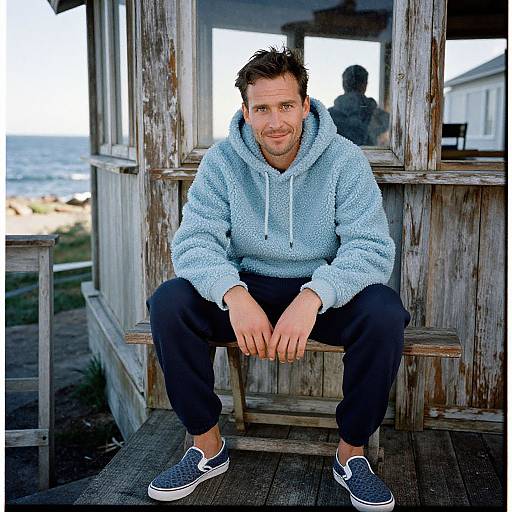 Photograph of a smiling, bearded man with short brown hair, wearing a light blue hoodie, black pants, and grey slip-on shoes, sitting