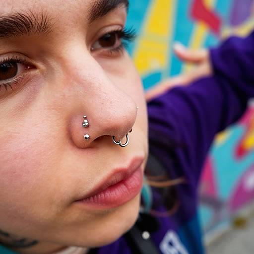 Close-up photograph of a young woman with light skin, thick eyebrows, and nose piercings, wearing a dark jacket, against a colorful graffiti background