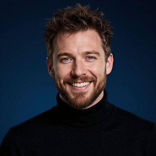 Photograph of a smiling Caucasian man with short, spiked brown hair, blue eyes, and a trimmed beard, wearing a black turtleneck, against