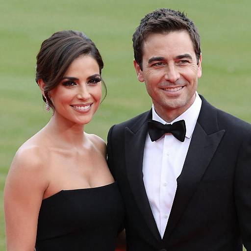 Photograph of a smiling couple in formal black attire; woman in strapless black dress, man in black tuxedo with bow tie, standing against