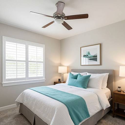 Bright bedroom photograph: white walls, gray bed skirt, teal blanket and pillows, white sheets, ceiling fan, two bedside lamps, framed landscape painting,