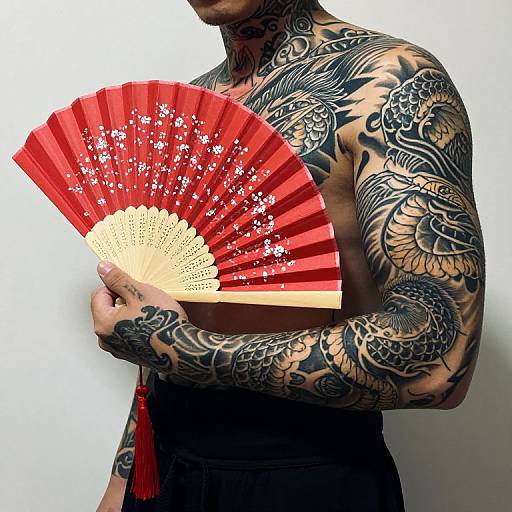 Tattooed shirtless man holding red floral fan with white blossoms, black pants, intricate dragon and floral tattoos, white background. Photograph.