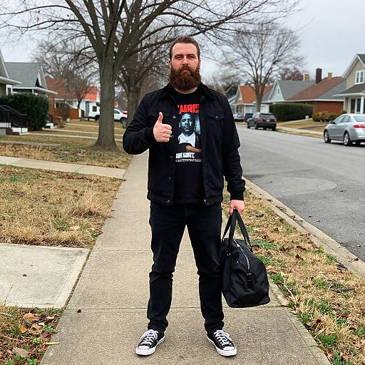 Photograph of a bearded man with a full beard, wearing a black jacket, graphic t-shirt, and black pants, giving a thumbs-up,