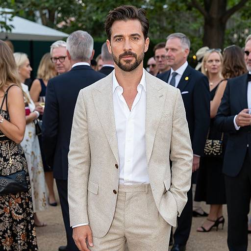 Photograph of a handsome bearded man with dark hair, wearing a light beige suit and white shirt, standing in front of a group of elegantly