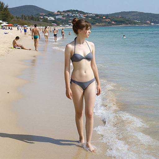 Photograph of a young woman in a gray bikini walking along a sunny, sandy beach with clear blue water and distant hills. Other beachgoers are