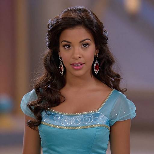 Photograph of a young woman with dark curly hair, wearing a blue, sparkling, short-sleeved dress and red diamond earrings, smiling against a