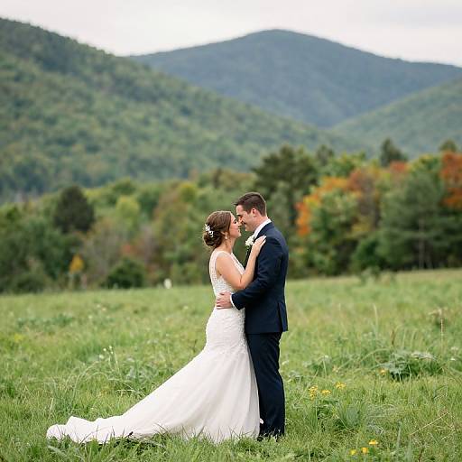 Photograph of a bride in a white strapless gown and groom in a black suit, kissing in a grassy field with forested mountains in the