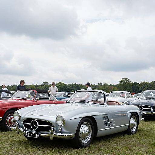 Photograph of a classic car show featuring a silver Mercedes-Benz convertible in the foreground, with red and gray cars behind, and several people standing under a