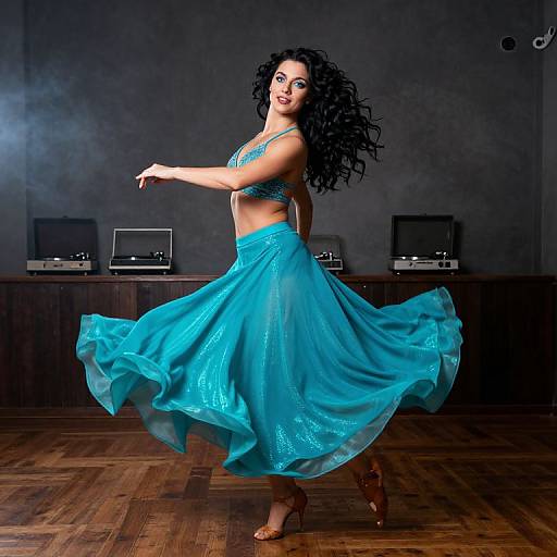 Photograph of a curly-haired woman in a blue belly dance outfit, mid-spin, against a dark, smoky background with wooden floor and stereo equipment