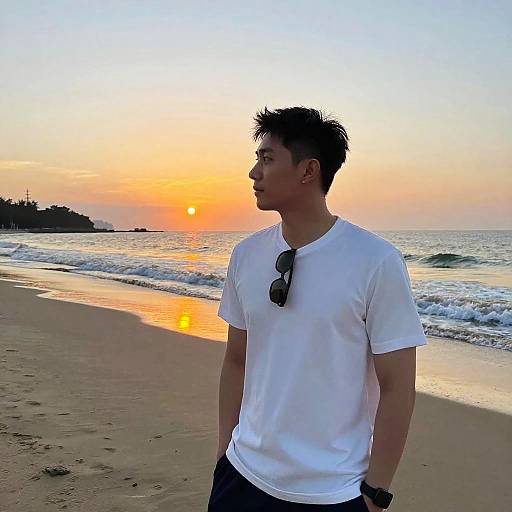 Photograph of a young Asian man with short black hair, wearing a white t-shirt and black pants, standing on a sandy beach at sunset, with