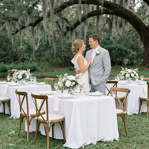 Photograph of a bride in a white gown and groom in a gray suit standing in an outdoor garden, surrounded by white-linen tables with floral center