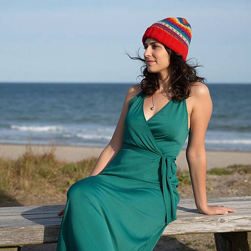 Photograph of a smiling woman with curly black hair, wearing a red knit beanie and green dress, sitting on a beach wooden bench, ocean and