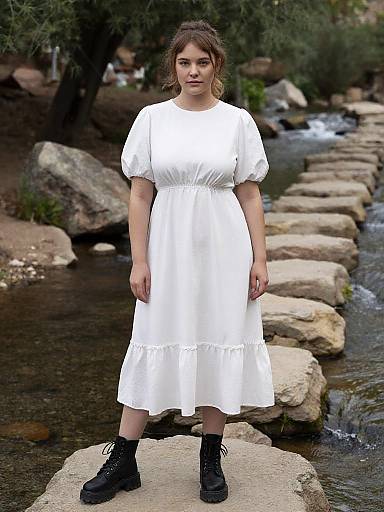 Photograph of a young woman with fair skin and brown hair in a white, short-sleeved, knee-length dress and black boots, standing on