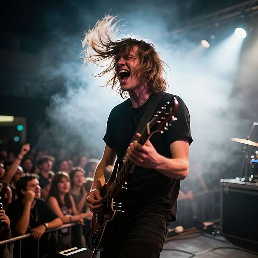 Photograph of a male rock musician with long brown hair, black t-shirt, playing electric guitar, mid-performance, stage lights, and cheering crowd in