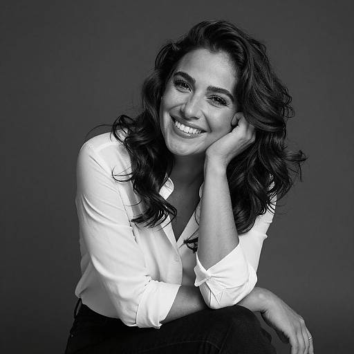 Black-and-white photograph of a smiling woman with wavy dark hair, wearing a white button-up shirt, resting her cheek on her hand. Dark background