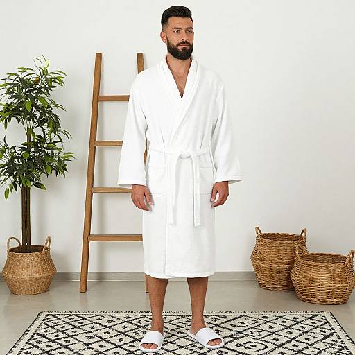 Photograph of a bearded man in a white bathrobe, standing in front of a wooden ladder, with wicker baskets and a potted plant