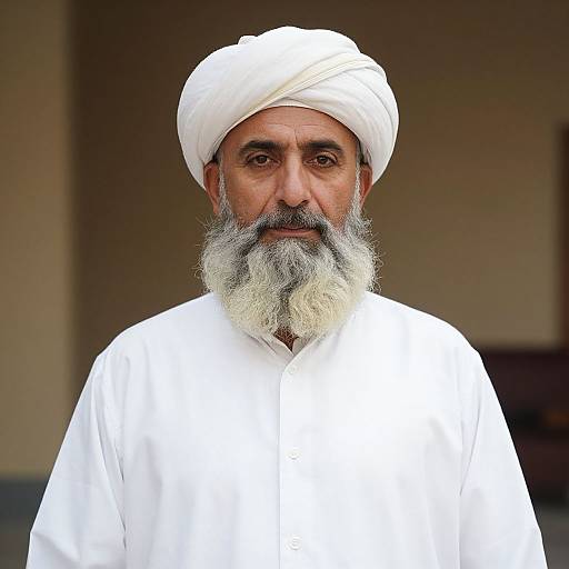 Photograph of an elderly Middle Eastern man with a white turban, white beard, and white robe, standing against a blurred beige background.