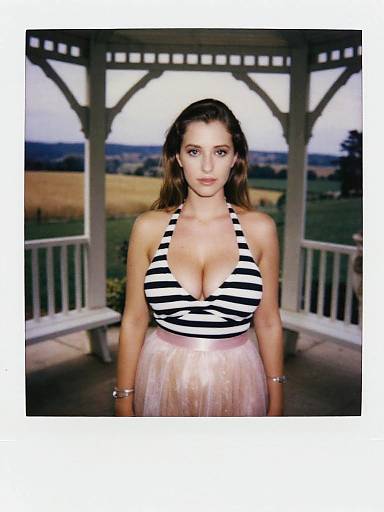 Photograph of a young woman with long brown hair, wearing a black-and-white striped halter top and pink tulle skirt, standing on a porch