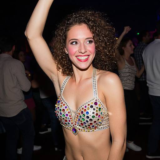 Photograph of a smiling, curly-haired woman with light skin wearing a sparkling, multicolored bra, raising her arm in a dark, crowded club
