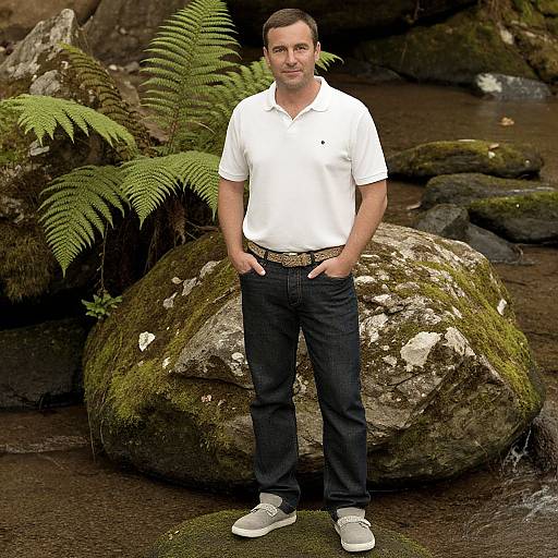 Photograph of a smiling man in a white polo, dark jeans, and gray sneakers standing in front of moss-covered rocks and ferns by a stream