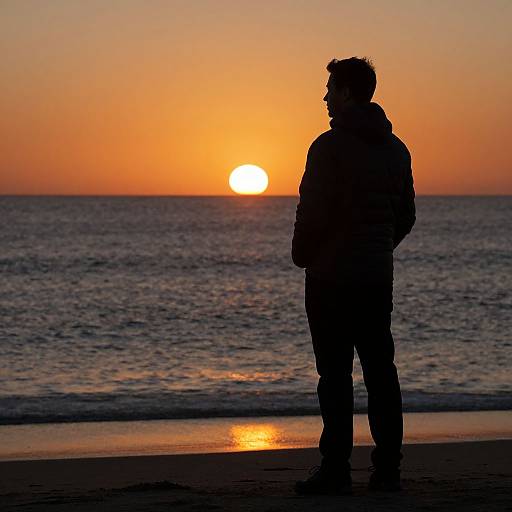 Silhouetted person standing on beach at sunset, with the sun partially visible over calm ocean, casting orange and orange-blue hues. Photograph.