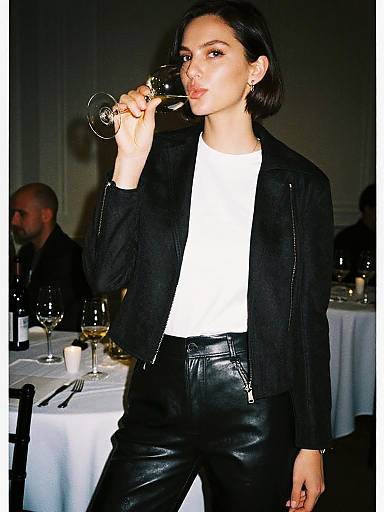 Photograph of a stylish woman with short dark hair, black leather pants, white shirt, and black jacket, sipping wine at a dimly lit