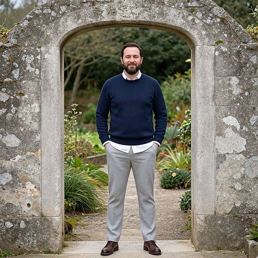Photograph of a bearded man with short dark hair, wearing a black sweater, white shirt, gray pants, and brown shoes, standing in front
