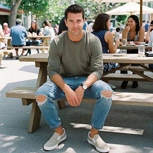Photograph of a muscular man with short dark hair, wearing a gray long-sleeve shirt, ripped blue jeans, and white sneakers, sitting on