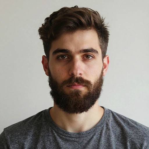 Photograph of a young man with a full beard, dark brown hair, and brown eyes, wearing a gray t-shirt, against a plain white background