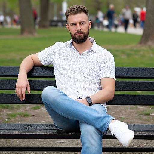 Photograph of a bearded, brown-haired man with a muscular build, wearing a white patterned shirt, blue jeans, and white sneakers, sitting