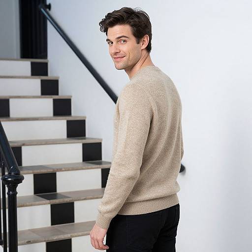Photograph of a smiling, medium-build man with dark hair, wearing a beige sweater and black pants, standing on a black-and-white staircase.