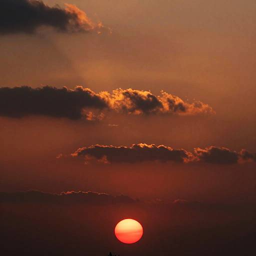 Photograph of a vibrant sunset with a glowing red sun, surrounded by dark orange and black clouds, casting dramatic shadows in the sky.