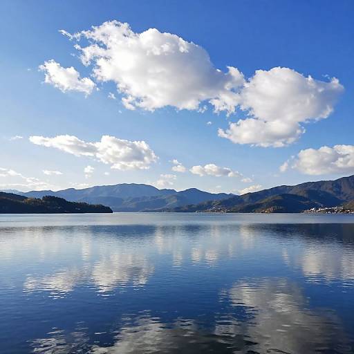 Photograph of a serene lake with calm, reflective water, surrounded by blue mountains under a bright, cloud-filled sky.