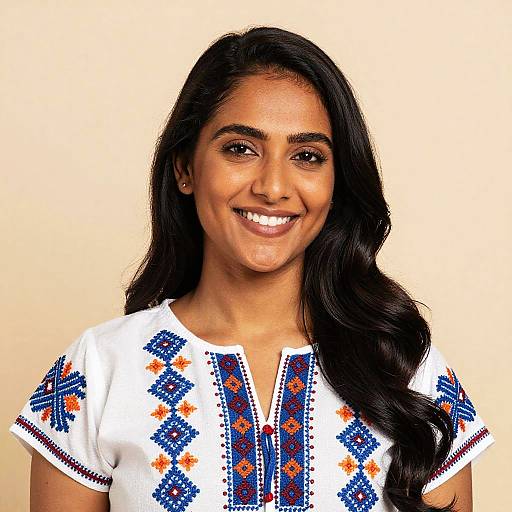 Photograph of a smiling Indian woman with long black hair, wearing a white dress with blue and orange geometric embroidery, against a plain beige background.