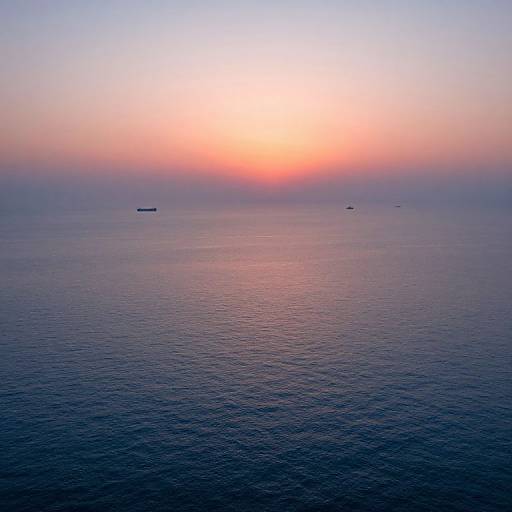 Photograph of a serene ocean at sunset, featuring a gradient sky from bright orange to soft pink, with small ships dotting the calm, darkening
