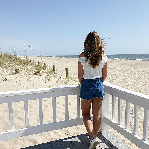 Photograph of a woman with long brown hair, wearing a white off-shoulder top and blue denim shorts, standing on a white wooden beachboard