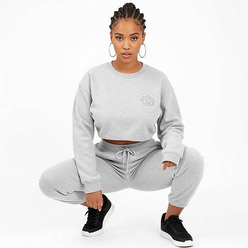 Photograph of a young Black woman with braided hair, wearing a gray sweatshirt, matching sweatpants, and black sneakers, squatting against a