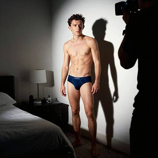 Photograph of a shirtless, slim, young man with curly hair wearing dark blue briefs, standing in a dimly lit bedroom, shadowed