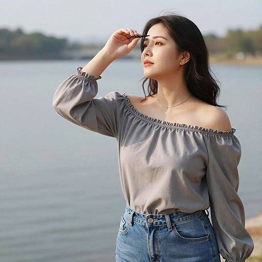 Photograph of an Asian woman with long black hair, wearing an off-the-shoulder gray blouse and high-waisted blue jeans, standing by
