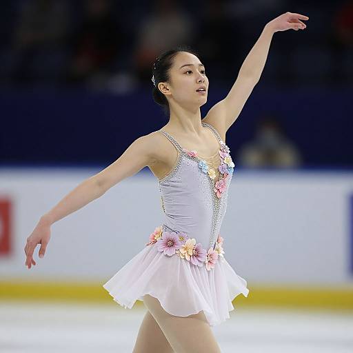 Photograph of an Asian female ice skater in a white, flower-adorned dress, extending her arm gracefully on an ice rink.