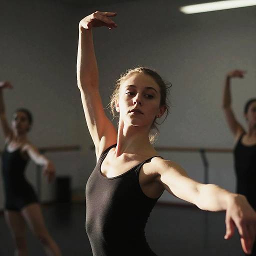 Photograph of a focused ballerina in a black leotard, arm raised, illuminated by overhead light, with two blurred dancers in the background