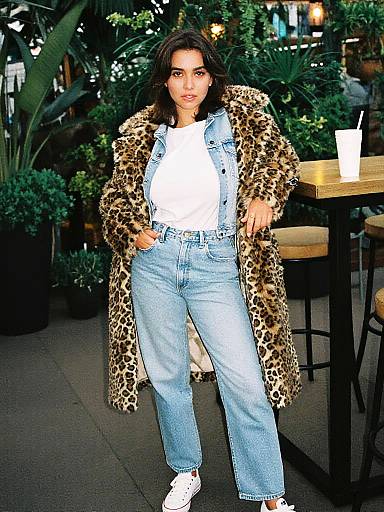 Photograph of a woman with dark hair, wearing a leopard print coat, white shirt, blue jeans, and white sneakers, standing in a lush,
