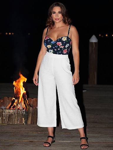 Photograph of a woman with curly brown hair, wearing a floral black top and white high-waisted pants, standing in front of a burning fire