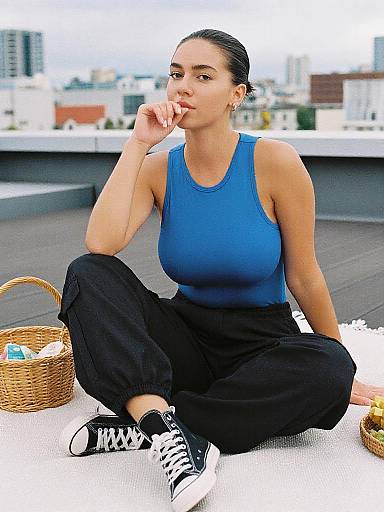 Photograph of a fit woman with medium brown skin, dark hair in a bun, wearing a blue tank top and black pants, sitting on a rooftop