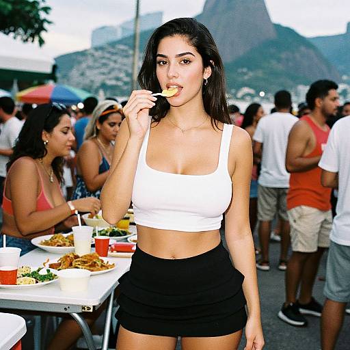 Photograph of a young woman with long black hair, wearing a white crop top and black shorts, eating a bite outdoors, surrounded by a diverse group