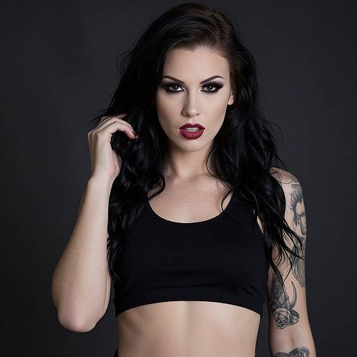 Photograph of a pale-skinned woman with long black hair, bold red lipstick, black crop top, tattooed arm, dark background.