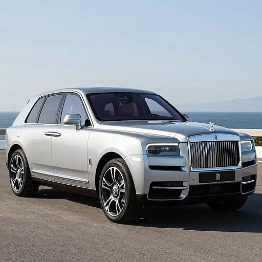 Photograph of a sleek, silver Rolls-Royce Wraith luxury sedan parked on a sunlit, coastal road with clear blue sky and ocean in