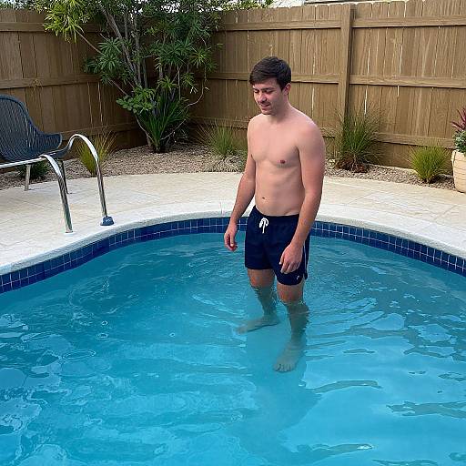Photograph of a young, shirtless man with short brown hair, wearing black swim trunks, standing in a backyard pool. Wooden fence and plants