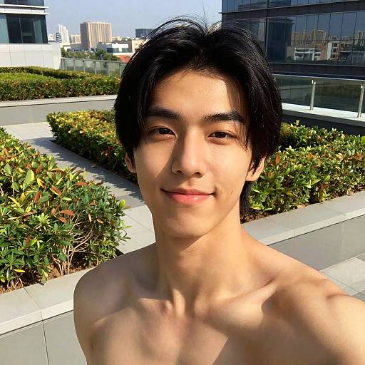 Photograph of a young Asian man with medium-length black hair, smiling, shirtless, standing outdoors in a modern urban garden with green bushes and glass